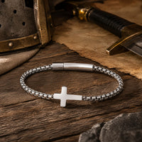 Bracelet du Pacte Sacré en acier inoxydable dans un décor médiéval avec casque, épée, parchemin et lumière chaude