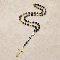 Chapelet de l’Ordre Sacré en acier inoxydable avec perles noires et dorées sur fond pierre médiévale claire.