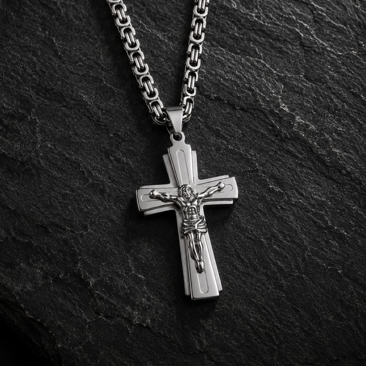 Variante argentée du Collier Croix du Christ en acier inoxydable sur fond anthracite texturé.