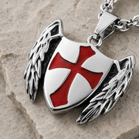 Gros plan du Collier du Gardien Croisé en acier inoxydable avec motif bouclier ailé et croix rouge sur fond pierre médiévale claire