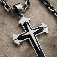 Gros plan de la croix noire et argentée du Collier du Gardien du Temple en acier inoxydable sur pierre médiévale claire.