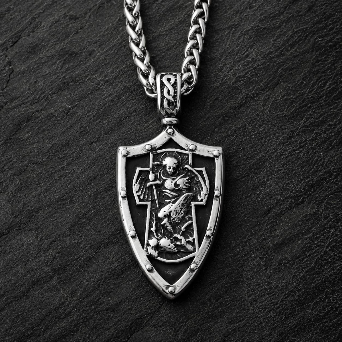 Collier Gloire de l’Archange en acier inoxydable avec pendentif bouclier argenté sur fond anthracite texturé.