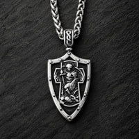 Collier Gloire de l’Archange en acier inoxydable avec pendentif bouclier argenté sur fond anthracite texturé.