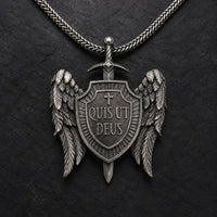 Collier Quis Ut Deus en acier inoxydable avec pendentif bouclier ailé sur fond anthracite texturé.