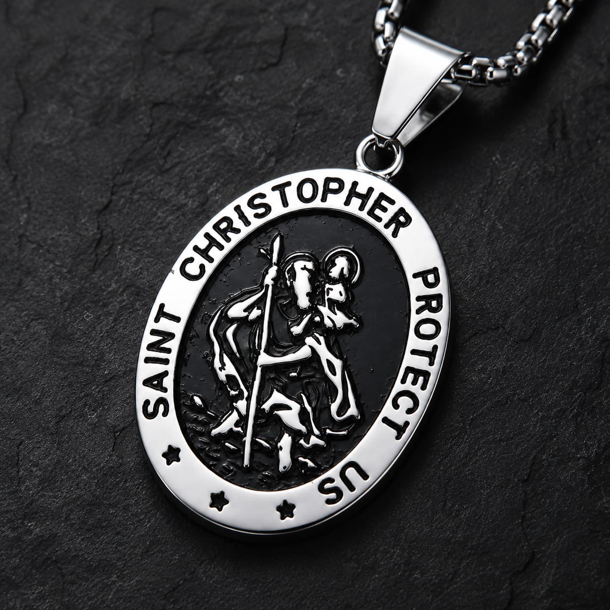 Gros plan du pendentif ovale du collier Saint Christophe Protecteur en acier inoxydable sur fond anthracite texturé.