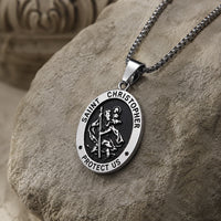 Vue de trois quarts du collier Saint Christophe Protecteur en acier inoxydable avec pendentif ovale argenté et noir sur pierre médiévale claire.