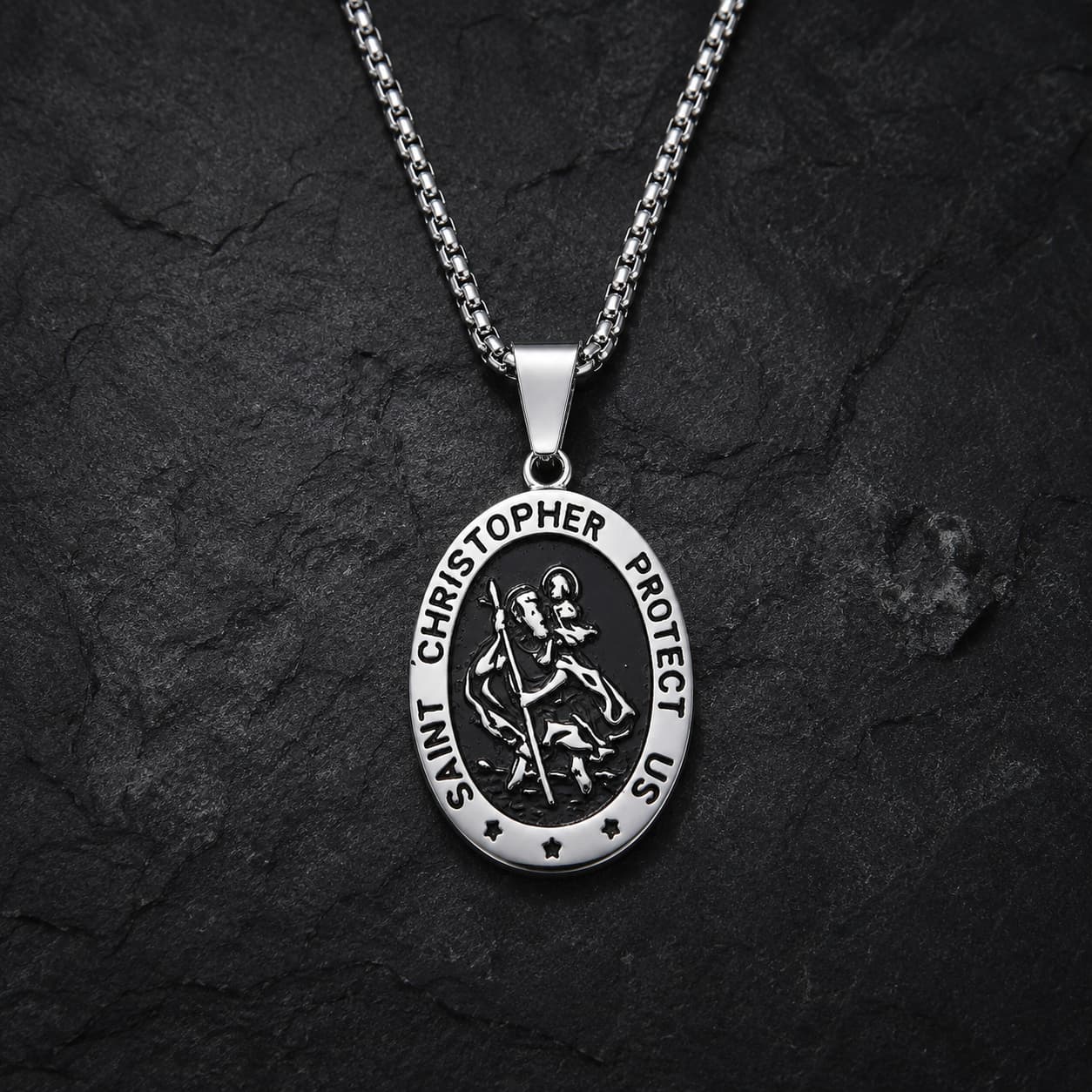 Collier Saint Christophe Protecteur en acier inoxydable avec pendentif ovale argenté et noir sur fond anthracite texturé.
