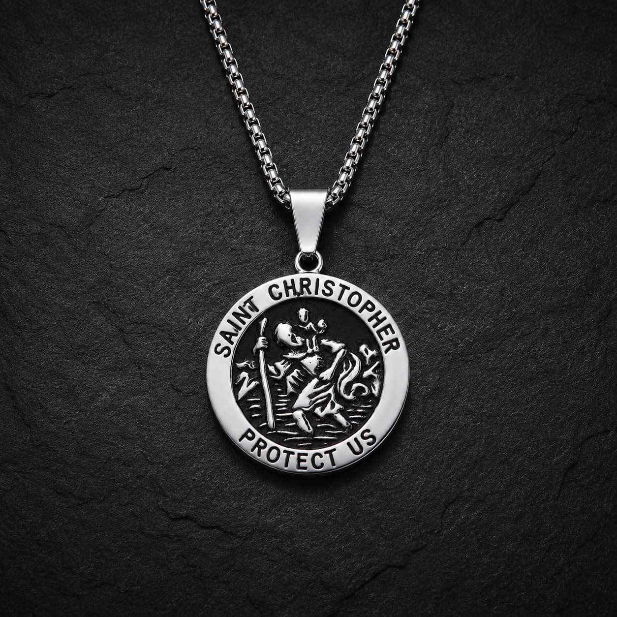 Variante ronde du collier Saint Christophe Protecteur en acier inoxydable avec finition argentée et noire sur fond anthracite texturé.