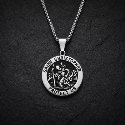 Variante ronde du collier Saint Christophe Protecteur en acier inoxydable avec finition argentée et noire sur fond anthracite texturé.