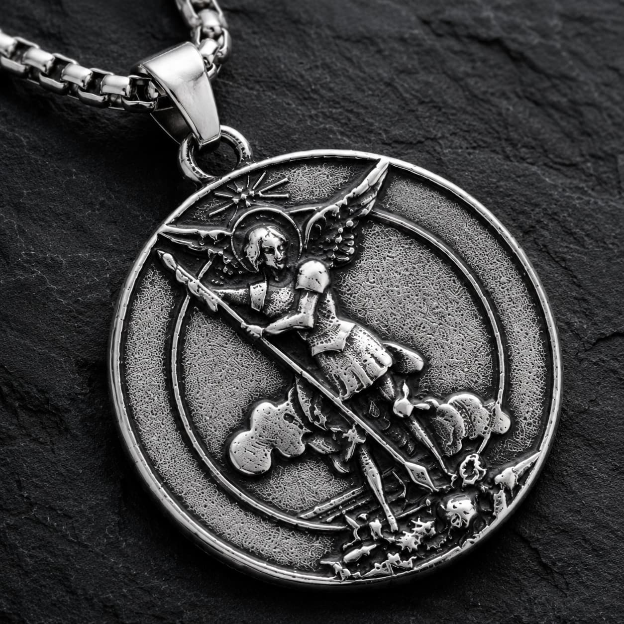 Gros plan du motif du Collier Saint Michel Protecteur en acier inoxydable sur fond anthracite texturé ardoise sombre