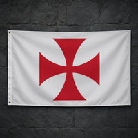 Drapeau blanc à croix pattée rouge en polyester accroché sur un mur de pierre sombre médiéval.