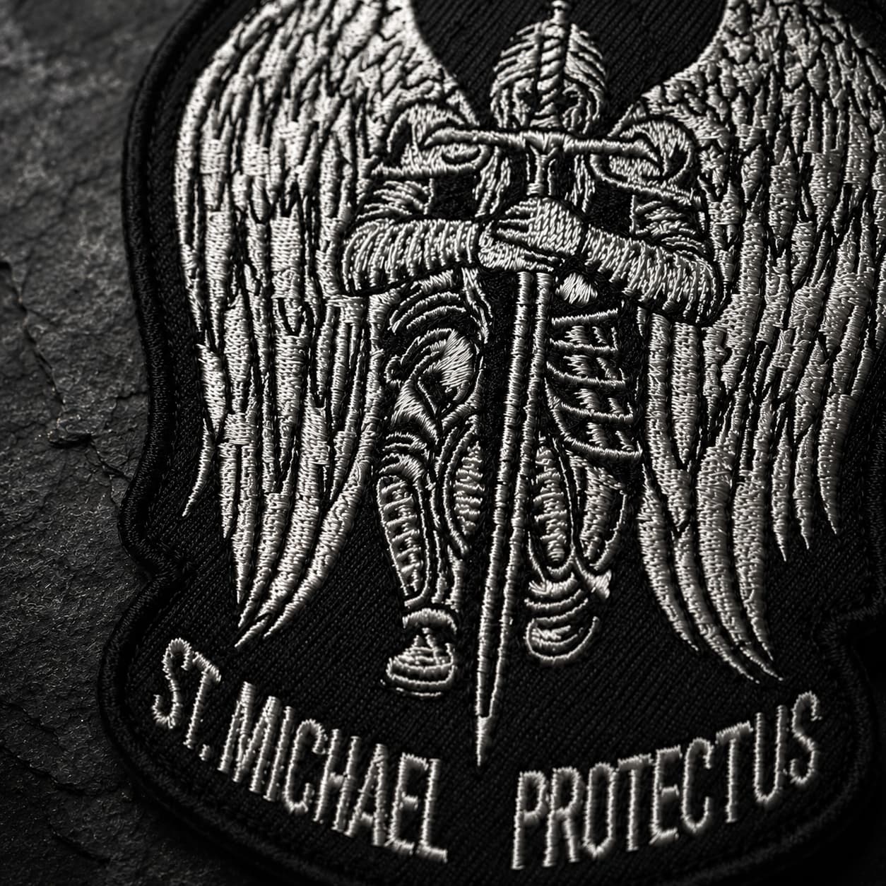Gros plan sur la broderie de l'Écusson de l'Archange Protecteur avec ailes, armure et épée.