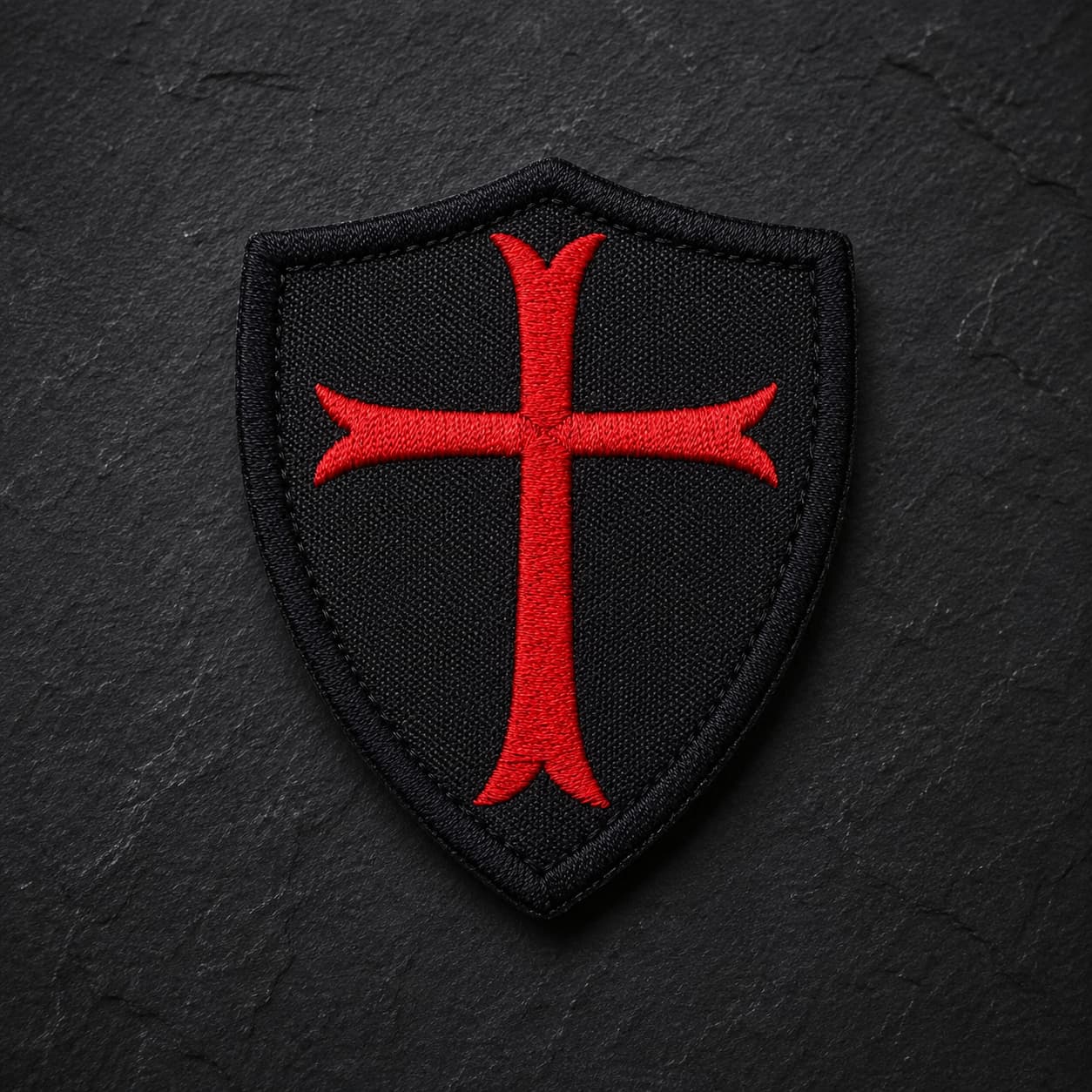 Écusson Chevalier Croisé en tissu brodé noir avec croix rouge sur fond ardoise sombre.