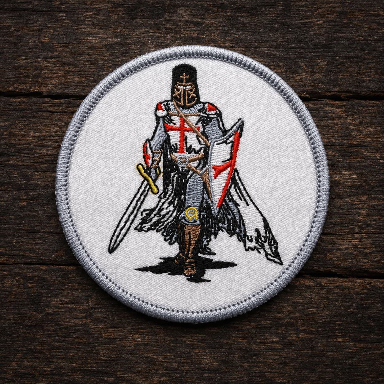 Variante ronde de l’Écusson Chevalier Templier en tissu brodé posée sur bois ancien sombre.