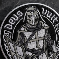 Gros plan sur la broderie de l’Écusson Deus Vult du Croisé en tissu brodé résistant sur fond ardoise sombre.