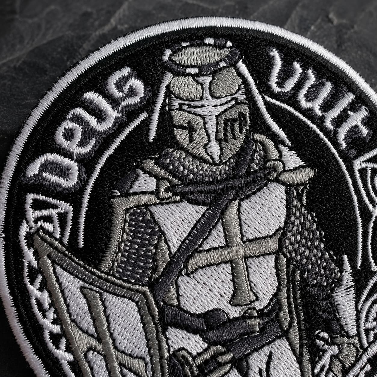 Gros plan sur la broderie de l’Écusson Deus Vult du Croisé en tissu brodé résistant sur fond ardoise sombre.