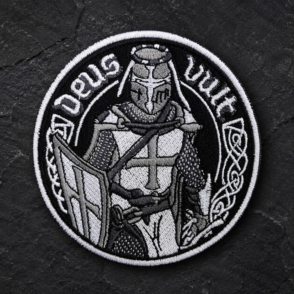 Écusson Deus Vult du Croisé en tissu brodé résistant sur fond de pierre sombre, avec chevalier templier en noir et blanc.