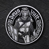 Écusson Deus Vult du Croisé en tissu brodé résistant sur fond de pierre sombre, avec chevalier templier en noir et blanc.