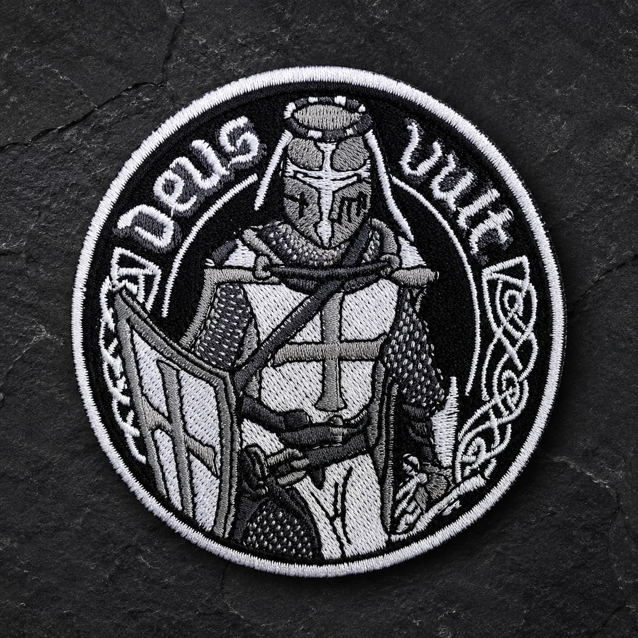 Écusson Deus Vult du Croisé en tissu brodé résistant sur fond de pierre sombre, avec chevalier templier en noir et blanc.