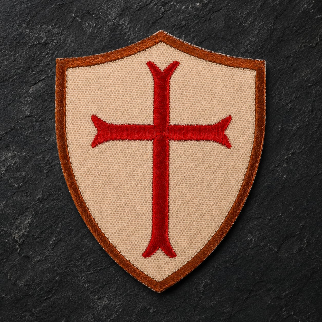 Écusson du Croisé en tissu brodé sur fond d’ardoise sombre, avec bouclier beige et croix rouge.