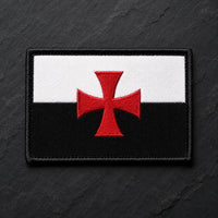 Ecusson du Devoir en tissu brodé noir et blanc avec croix rouge sur fond ardoise sombre.