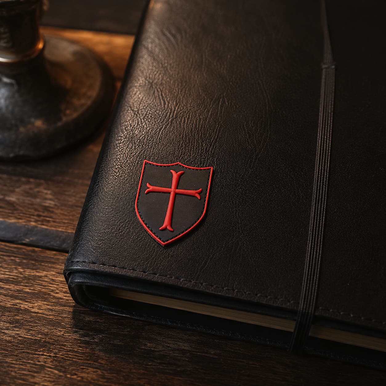 Écusson du Templier Royal en PVC appliqué sur un carnet en cuir noir dans une ambiance médiévale sombre.