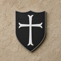 Écusson du Templier Royal en PVC noir avec croix blanche et bordure argentée sur fond de pierre claire.