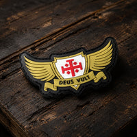 Patch Deus Vult du Croisé en PVC souple en vue inclinée sur bois ancien sombre, avec relief et bordure noire visibles.