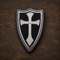 Variante noire du patch Écusson de la Lance du Templier en tissu brodé sur fond en bois ancien sombre.