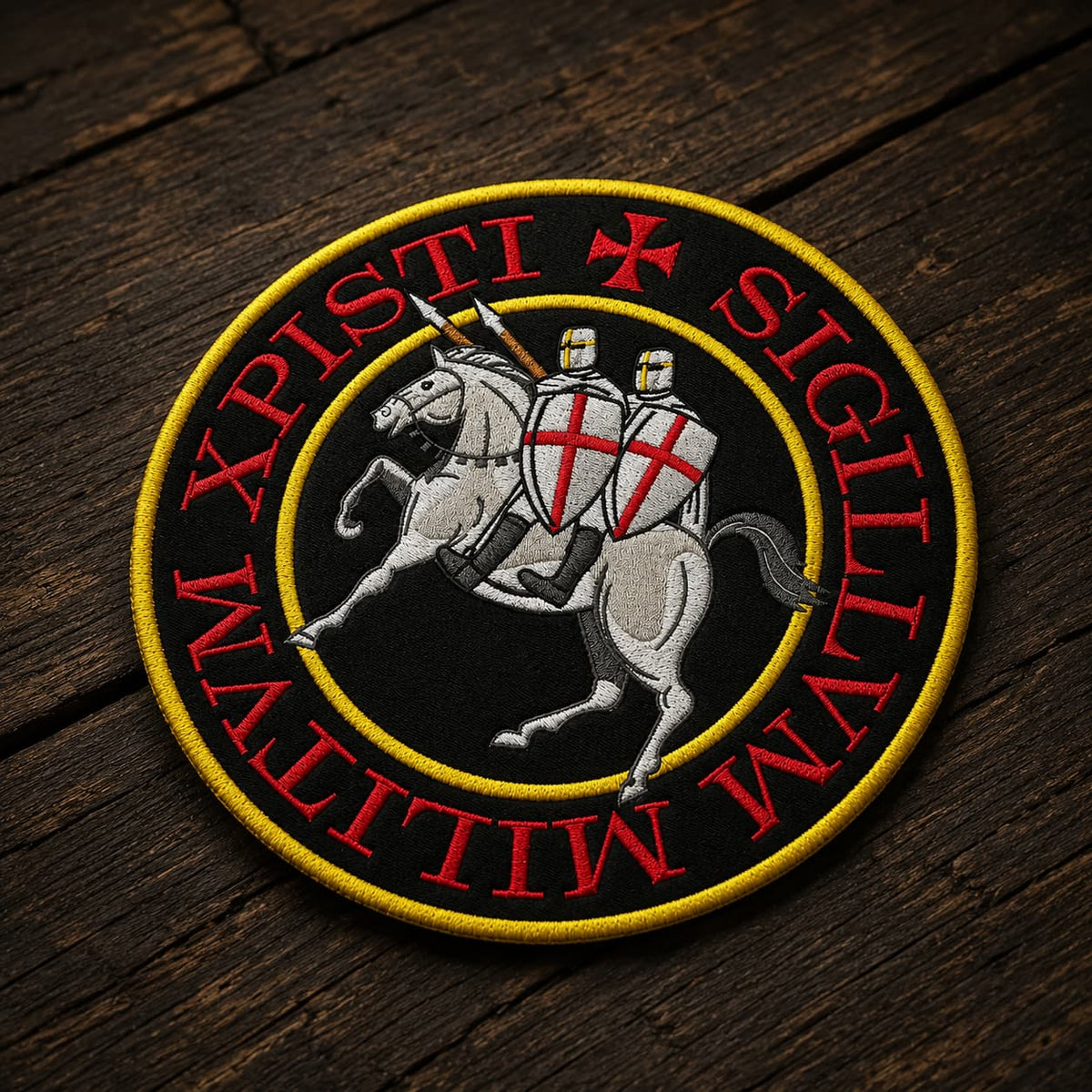 Patch Écusson Grand Sigillum Militum Xpisti en tissu brodé présenté en vue angle sur bois ancien, avec broderie rouge, noire, grise et jaune.