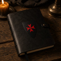 Sticker Bouclier Templier en vinyle adhésif appliqué sur un carnet en cuir noir dans une ambiance médiévale chaleureuse.